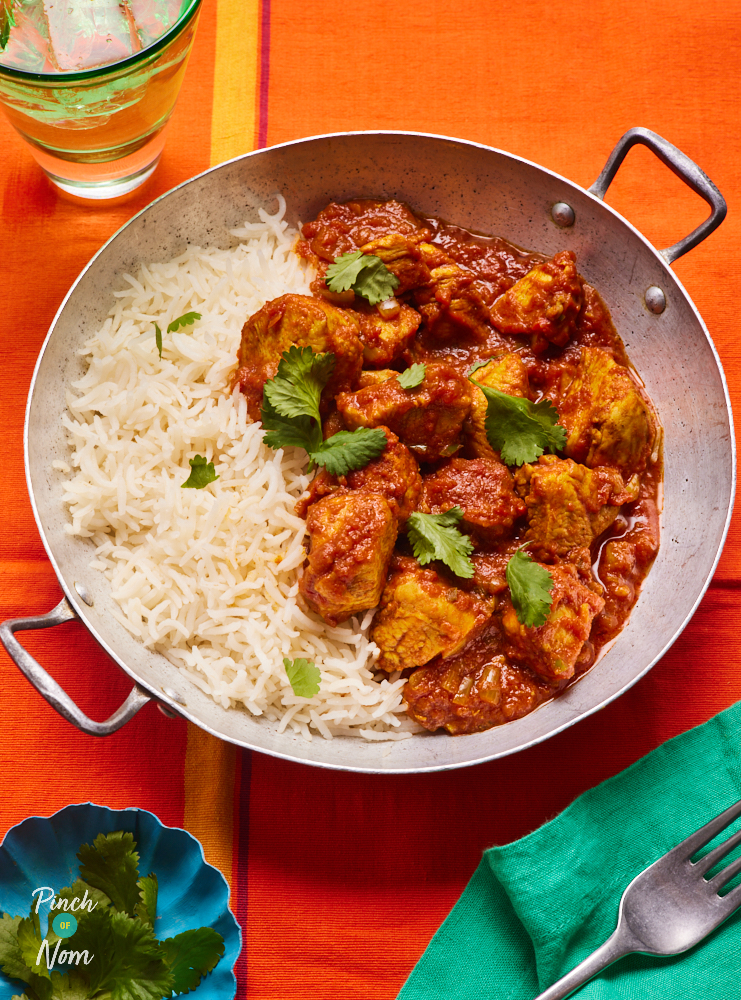 A table is set with a vibrant red tablecloth. In the centre, Pinch of Nom's Chicken Madras is served in a large silver, shallow pan with handles on each side. Half of the pan is filled with curry, and the other half is filled with fluffy white basmati rice. Coriander has been sprinkled on top.