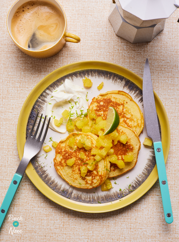 A table is set for breakfast, with a pot of coffee and a mug nearby. On a white plate with a yellow rim, 3 Piña Colada Pancakes are served, with a topping of pineapples, a slice of lime and a dollop of Greek yoghurt. A knife and fork with bright blue handles are resting on the plate.