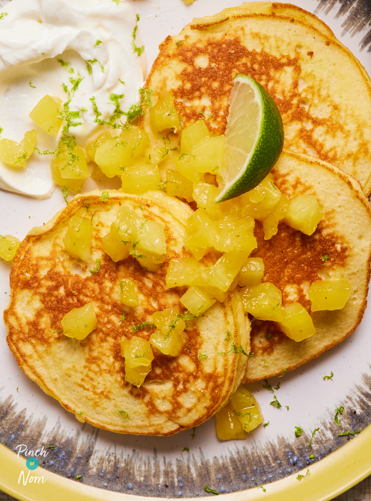 A close up shows Pinch of Nom's Piña Colada Pancakes served on a white plate with a yellow rim. The pancakes are topped with chopped pineapple, a wedge of lime and a dollop of creamy Greek yoghurt.