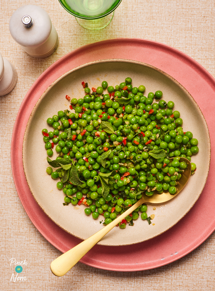 A table is set with a full plate of Pinch of Nom's Chilli Mint Peas on a pale pink set of crockery with a blush pink table mat. A gold spoon waits to tuck in.