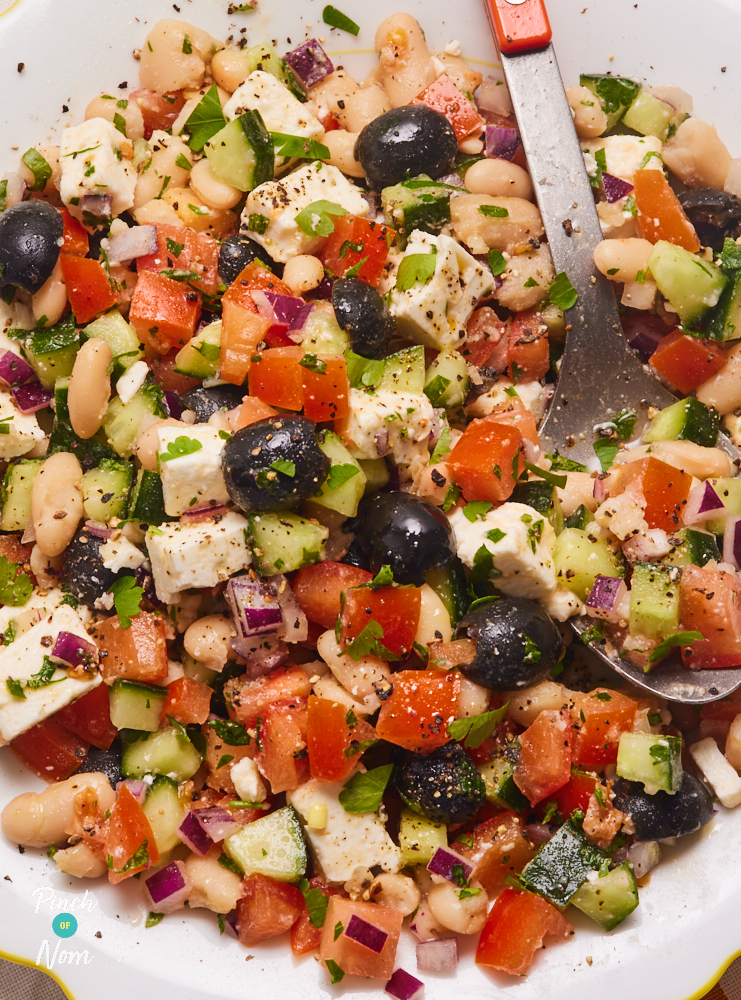 A close-up of Pinch of Nom's Greek-Style Bean Salad. It is a medley of chopped cucumber, tomatoes, red onion, black olives, feta cheese, parsley and cannellini beans. A metal serving spoon is resting in the centre, ready to dish up a portion.