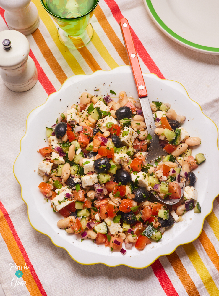 Pinch of Nom's Greek-Style Bean Salad is served on a large white plate with a yellow scalloped edge. The table is set with a colourful striped tablecloth, salt and pepper shakers and a glass of water. A metal serving spoon rests in the salad, ready to dish it up onto a plate that waits nearby.