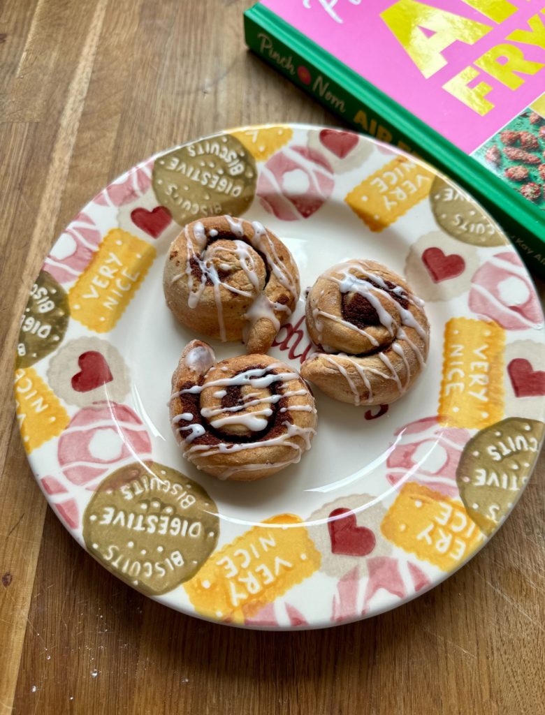 A plate is filled with 3 of Pinch of Nom's Cinnamon Buns from the Air Fryer cookbook, drizzled with a sticky-sweet glaze.