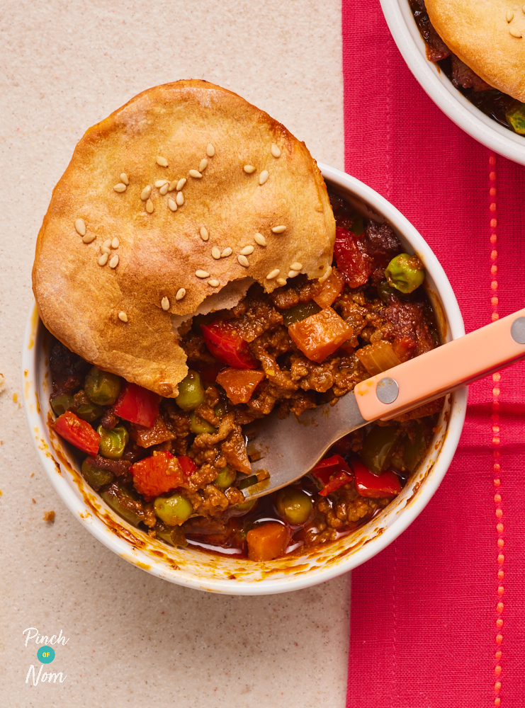 A close-up of one of Pinch of Nom's Keema Naan Pies, served on a table laid with a red cloth. The ramekin is filled with keema mixture, topped with a golden, homemade naan. A fork rests in the ramekin, and a bite has been taken from the naan pie topping.