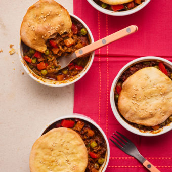 Pinch of Nom's Keema Naan Pies are served on a table laid with a red cloth. 4 ramekins are filled with keema mixture, topped with golden, homemade naans. A fork rests in one of the ramekins, and a bite has been taken from the naan pie topping.