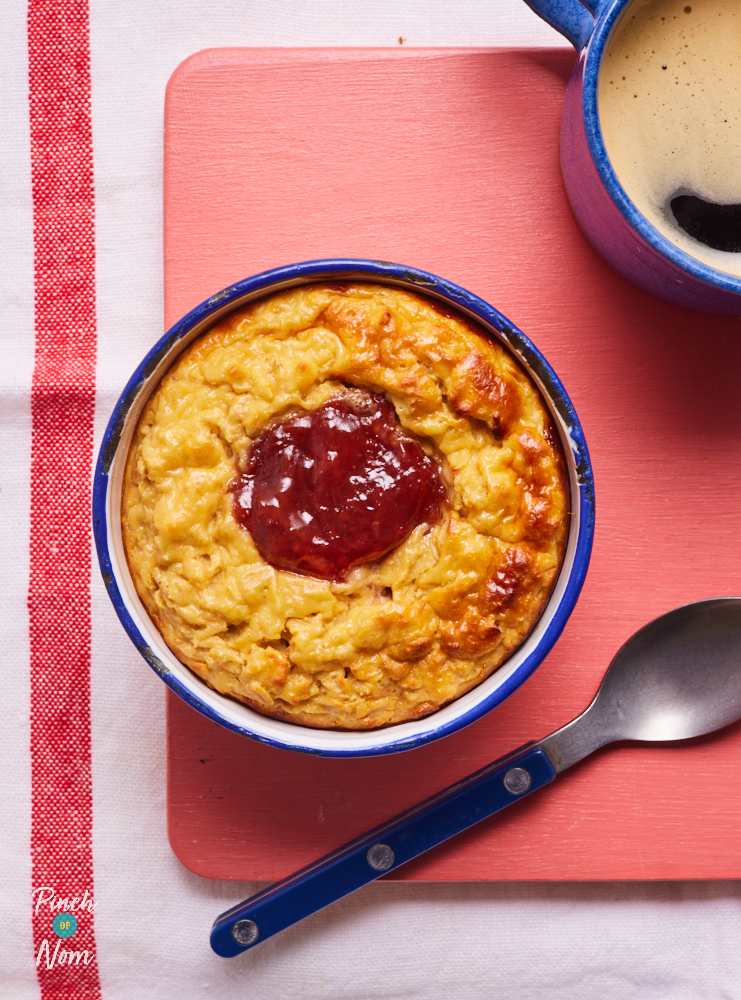 Pinch of Nom's Peanut Butter and Jelly Baked Oats are served for breakfast, alongside a cup of coffee. The oats have baked until golden on top, and strawberry jam is bubbling up in the middle.