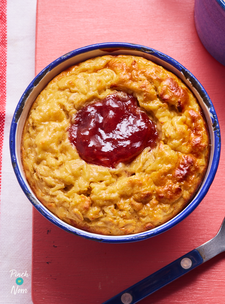 A close-up photo shows Pinch of Nom's Peanut Butter and Jelly Baked Oats served for breakfast. The oats have baked until golden on top, and strawberry jam is bubbling up in the middle.