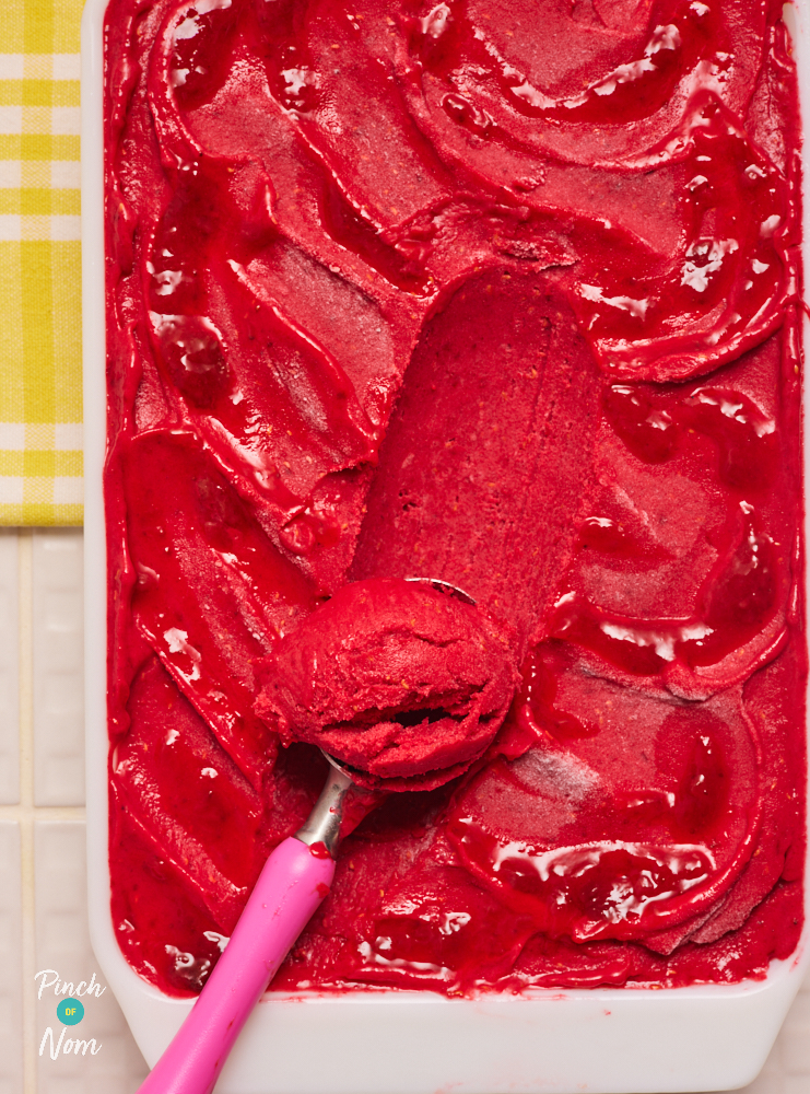 A close-up photo shows Pinch of Nom's low-calorie Raspberry Banana Ice Cream served in a large rectangular dish. An ice cream scoop with a pink handle rests in the centre of the dish, with a scoop of ice cream waiting to be dished up.