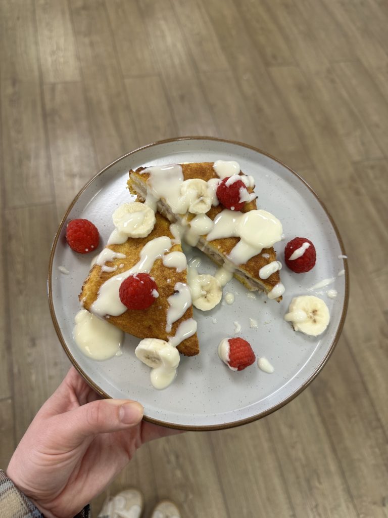 A plate of Pinch of Nom's Banana and Raspberry French Toast is served drizzled with added raspberries, freshly-sliced bananas and a generous drizzle of cream cheese and maple syrup sauce.