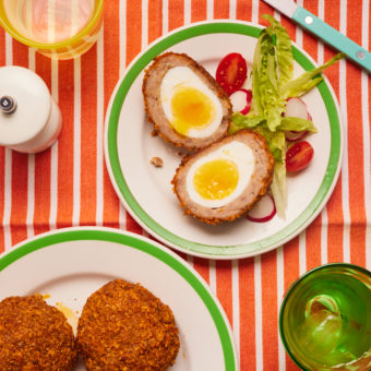 A table is laid with a red and white striped tablecloth. One of Pinch of Nom's Zinger Scotch Eggs is served with a side salad. The scotch egg has been cut in half, revealing the cross section of layers of freshly-boiled egg, sausage and crispy Zinger coating. On a plate nearby, the other Zinger Scotch Eggs are waiting to be served.