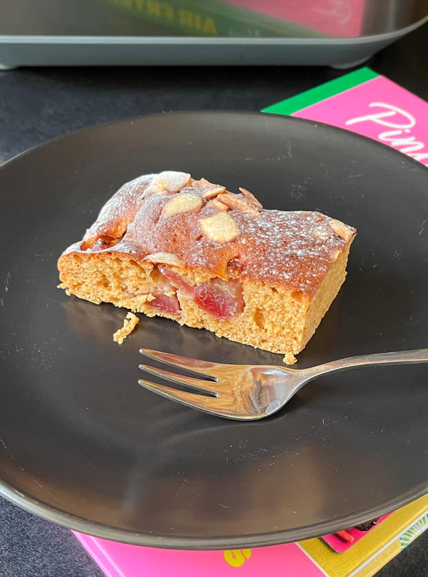 One slice of Pinch of Nom's Air Fryer Cherry Bakewell Slice recipe (from the Air Fryer cookbook) is plated up with a fork waiting to tuck in; cherries and almonds poke out from the sponge.