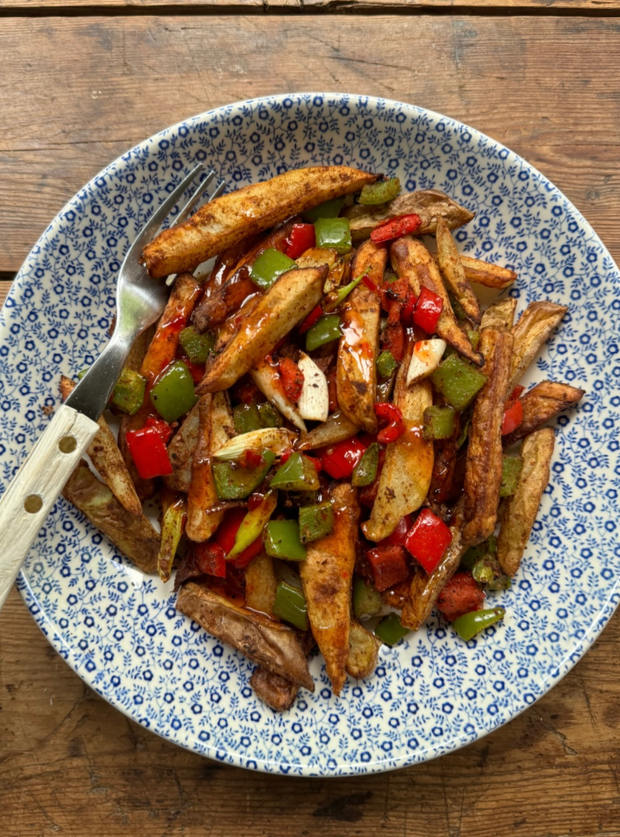 A portion of Pinch of Nom's Air Fryer Salt and Pepper Chips from the Air Fryer cookbook are served in a blue and white bowl with a fork waiting to dig in. They're mixed in with spices, chopped peppers and onions.