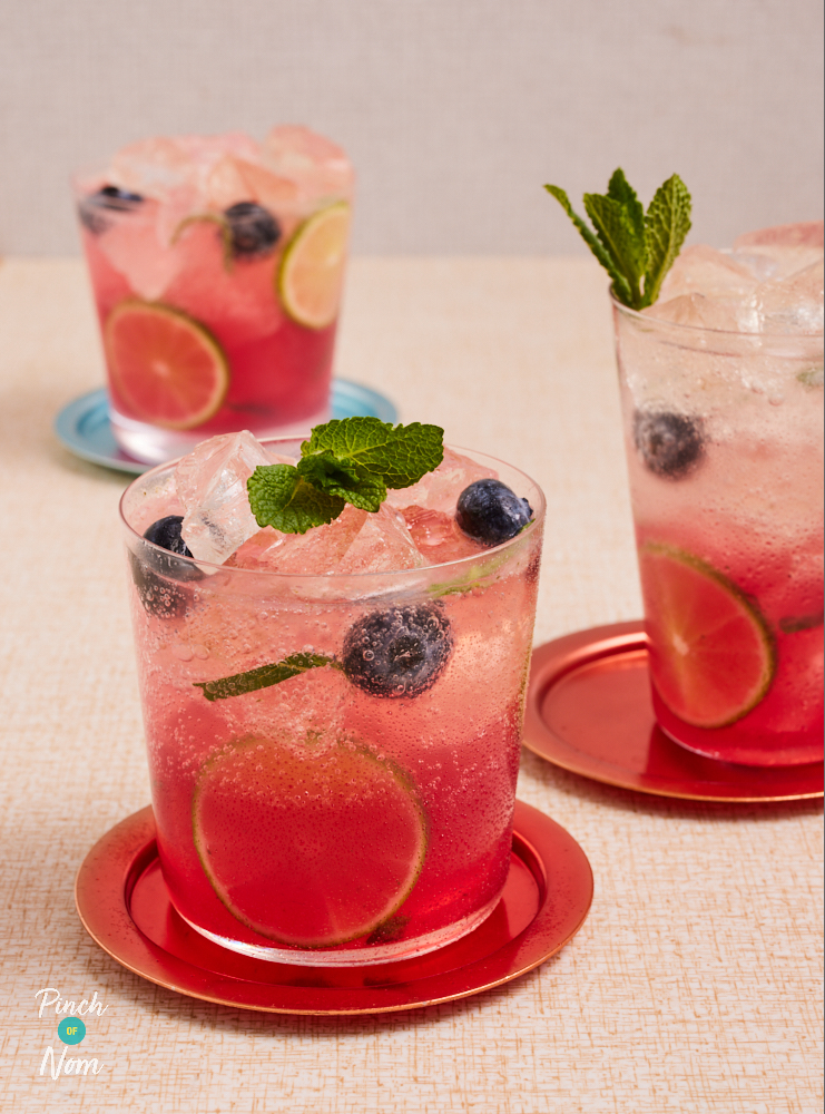 A table is set with three glasses of Pinch of Nom's slimming-friendly Blueberry Mojito cocktail. Each glass is topped with plenty of ice, fresh blueberries and a garnish of mint leaves.