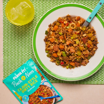 Flava It! X Pinch of Nom's Cajun-Style Dirty Rice is served on a white plate with a bright green rim. The plate is set on a green place mat, next to a sachet of the recipe mix and water in a bright yellow glass.