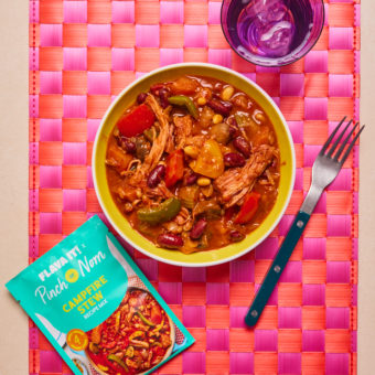 Pinch of Nom's Flava It! Campfire Stew is served in a bowl, on a pink and orange checked placemat. The table is set with a fork and a glass of water. The recipe mix sachet is placed next to the bowl of stew.