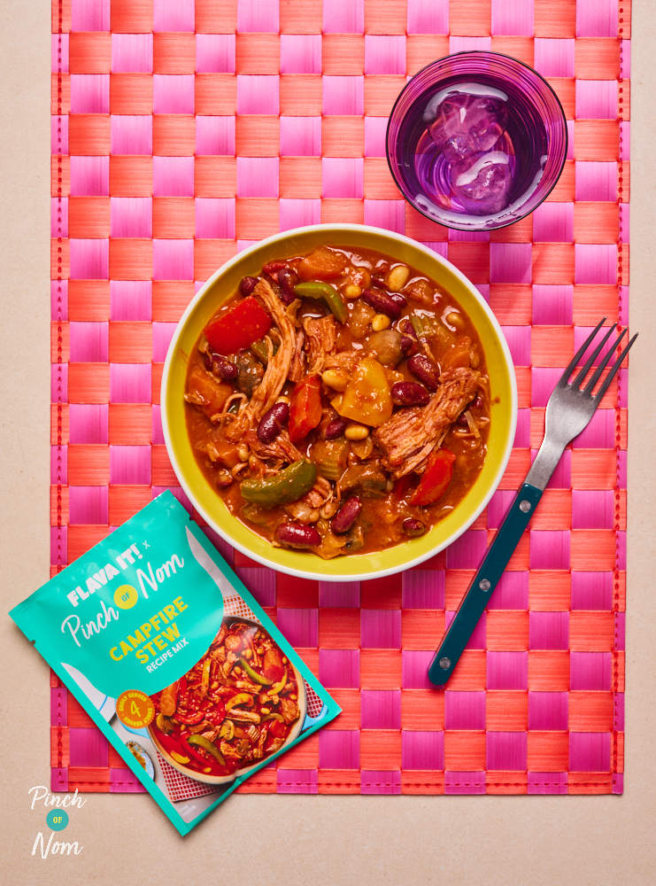 Pinch of Nom's Flava It! Campfire Stew is served in a bowl, on a pink and orange checked placemat. The table is set with a fork and a glass of water. The recipe mix sachet is placed next to the bowl of stew.