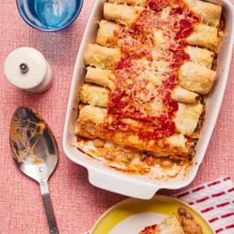 An oven dish is at the centre of a dinner table filled with Pinch of Nom's Creamy Chicken Enchiladas. They're topped with tomatoey sauce and a melty layer of reduced-fat Cheddar cheese.