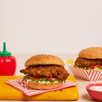 Pinch of Nom's Flava It! Crispy Southern Chicken is served in a burger bun, with salad. One burger is placed on a napkin, resting on a yellow chopping board. The other burger is in a red and white striped takeaway-style cardboard box in the background.