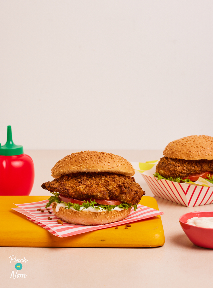 Pinch of Nom's Flava It! Crispy Southern Chicken is served in a burger bun, with salad. One burger is placed on a napkin, resting on a yellow chopping board. The other burger is in a red and white striped takeaway-style cardboard box in the background.