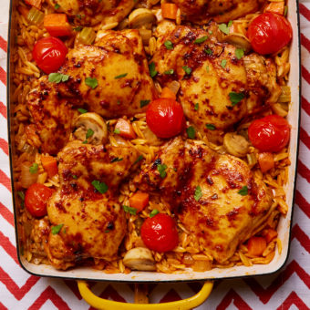 Pinch of Nom's Flava It! Mediterranean-Style Chicken Traybake is served in a large, rectangular oven dish, set on a board with a red and white zig zag pattern.