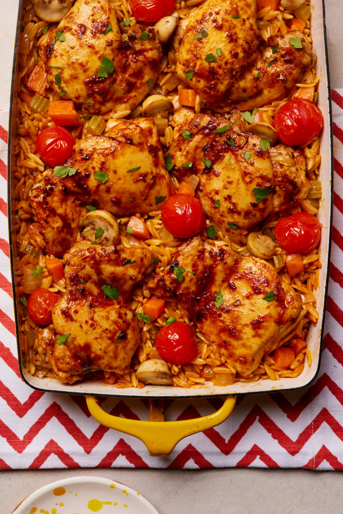 Pinch of Nom's Flava It! Mediterranean-Style Chicken Traybake is served in a large, rectangular oven dish, set on a board with a red and white zig zag pattern.