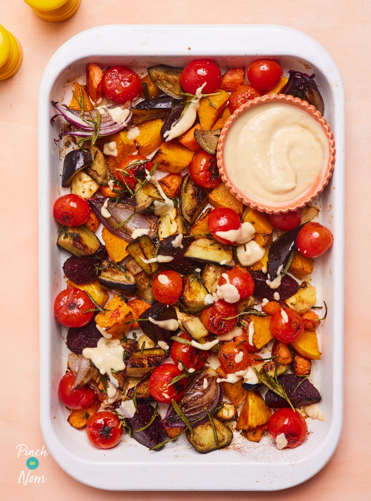 A white roasting tray is filled with Pinch of Nom's Autumn Vegetable Traybake; it includes a slightly crispy, golden array of cherry tomatoes, caramelised butternut squash, beetroot, red onion, aubergine and parsnips. Sprigs of rosemary and garlic are tossed through. A creamy sauce is drizzled on top, with a dipping sauce to the side in a small bowl, ready to tuck in.