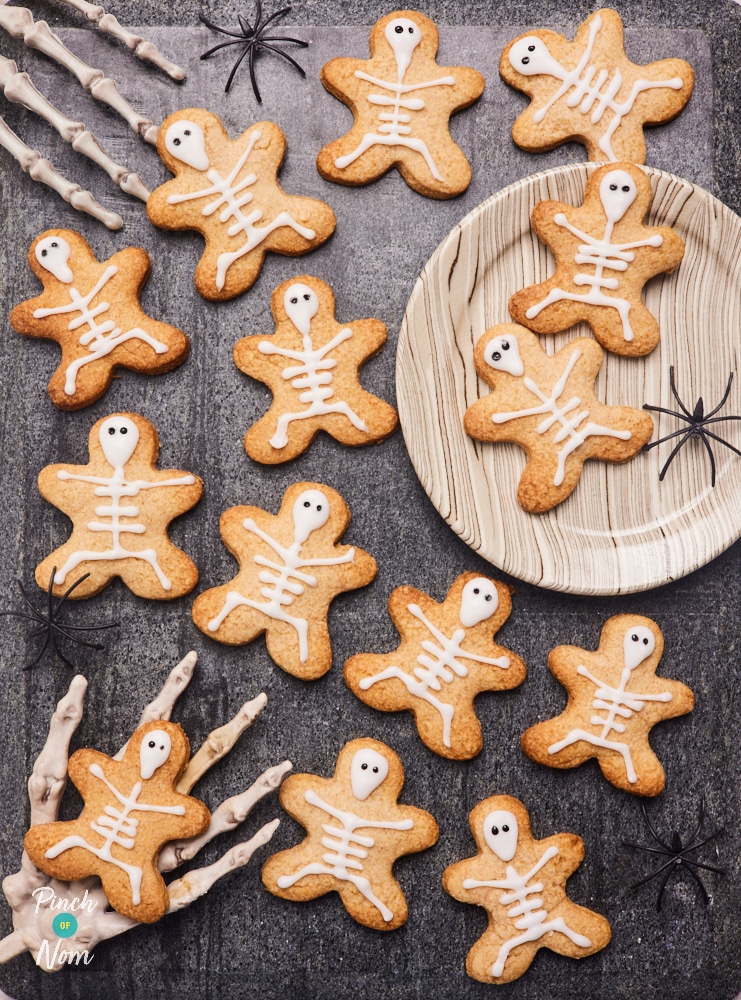 Pinch of Nom's spooky, slimming-friendly Halloween Skeleton Biscuits are scattered over a dark tabletop, with little spiders running by and a pair of skeleton hands waiting to tuck in.