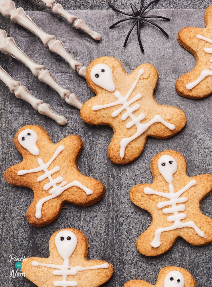 Pinch of Nom's spooky, slimming-friendly Halloween Skeleton Biscuits are scattered over a dark tabletop, with little spiders running by and a pair of skeleton hands waiting to tuck in.