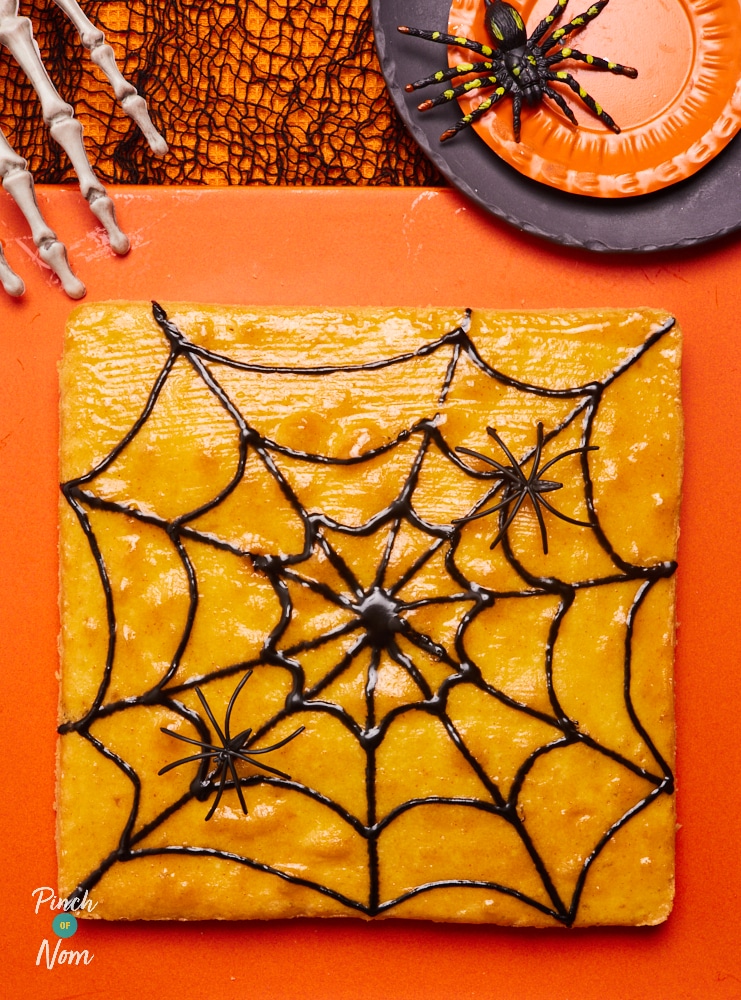 Pinch of Nom's Halloween Spooky Sponge is served on an orange board, surrounded by small plastic spider decorations. The golden sponge cake is decorated with a black spider web design. A skeleton hand decoration rests on the edge of the board, as if reaching for the cake.