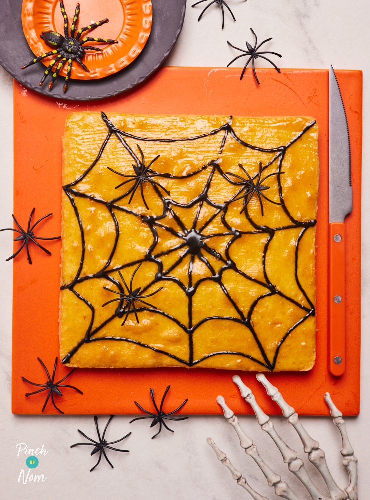 Pinch of Nom's Halloween Spooky Sponge is served on an orange board, surrounded by small plastic spider decorations. The golden sponge cake is decorated with a black spider web design, and an orange-handled knife is nearby, ready to slice and serve. A skeleton hand decoration rests on the edge of the board, as if reaching for the cake.