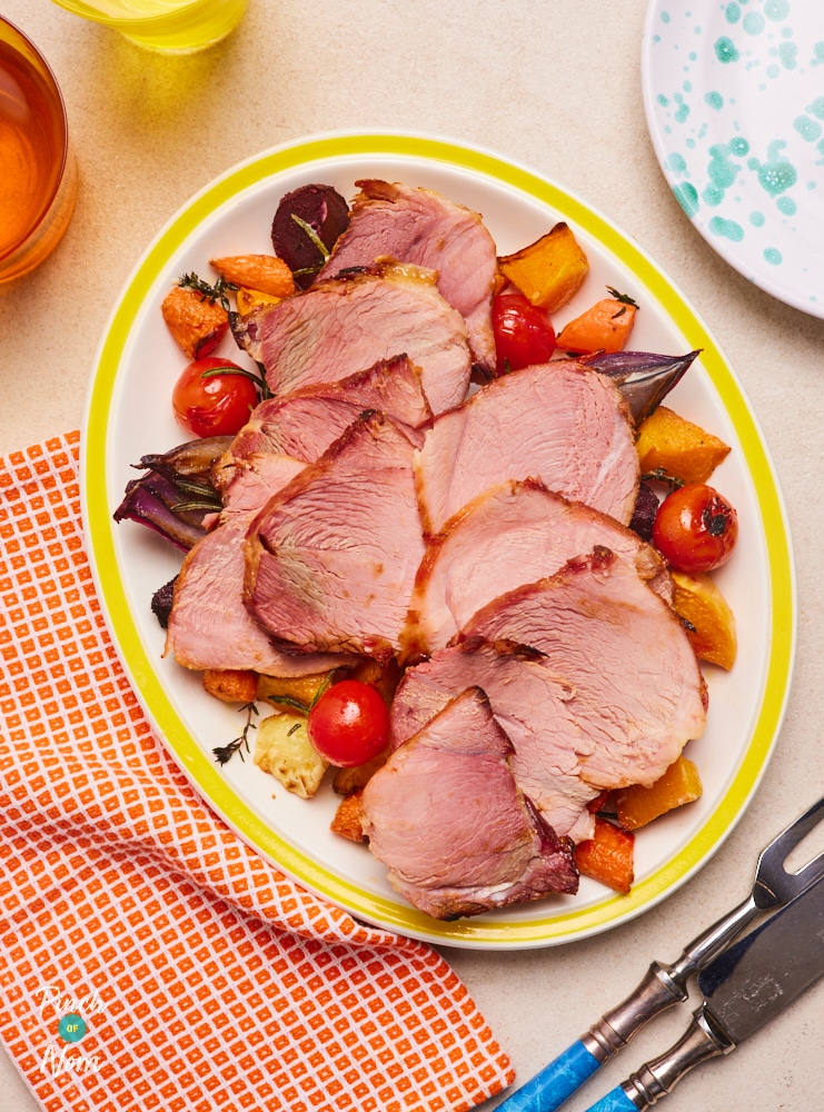 A large plate is filled with slices of Pinch of Nom's Honey-Glazed Ham, served on a colourful bed of browned, oven-roasted vegetables, including juicy cherry tomatoes, carrots and onions.