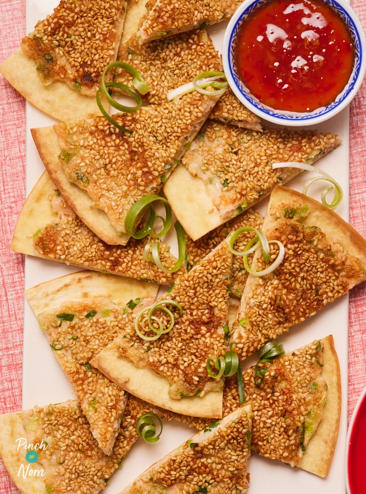 Pinch of Nom's Prawn Bites are served on a rectangular white board, set on a red tablecloth. The triangular bites are golden brown, and a pot of sweet chilli dipping sauce is on the board alongside them.