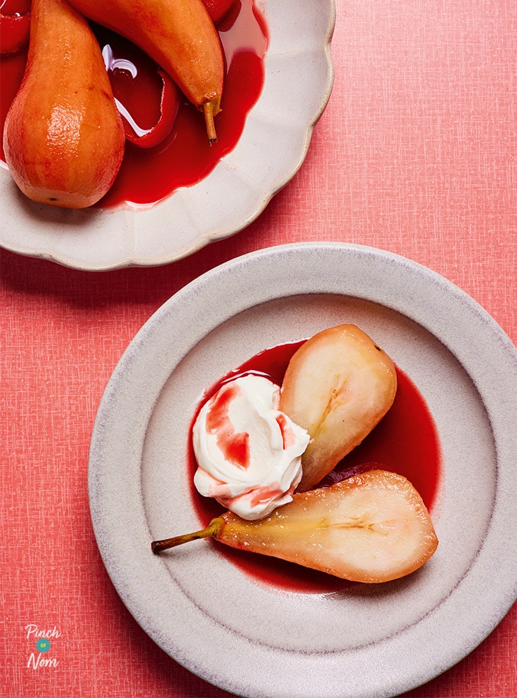 Pinch of Nom's slimming-friendly Cranberry Poached Pears are plated up on a festive table. The pears are served with a dollop of low-calorie cream, and coated in red-coloured syrup.