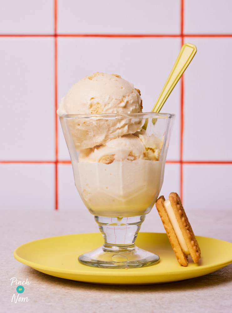 A dessert glass is filled with two large scoops of Pinch of Nom's Ninja Creami Custard Cream Ice Cream, with a custard cream biscuit served to the side, and a spoon poking out - ready to dig in.