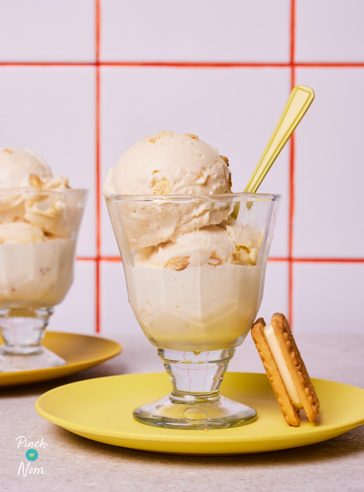 A dessert glass is filled with two large scoops of Pinch of Nom's Ninja Creami Custard Cream Ice Cream, with a custard cream biscuit served to the side, and a spoon poking out - ready to dig in.