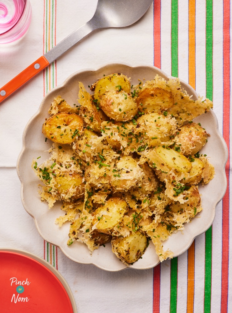 Pinch of Nom's Parmesan Potatoes are served in a white, scalloped-edged dish, on a table laid with a striped cloth, next to a red handled serving spoon. The potatoes are golden brown, with a crispy cheese crust.