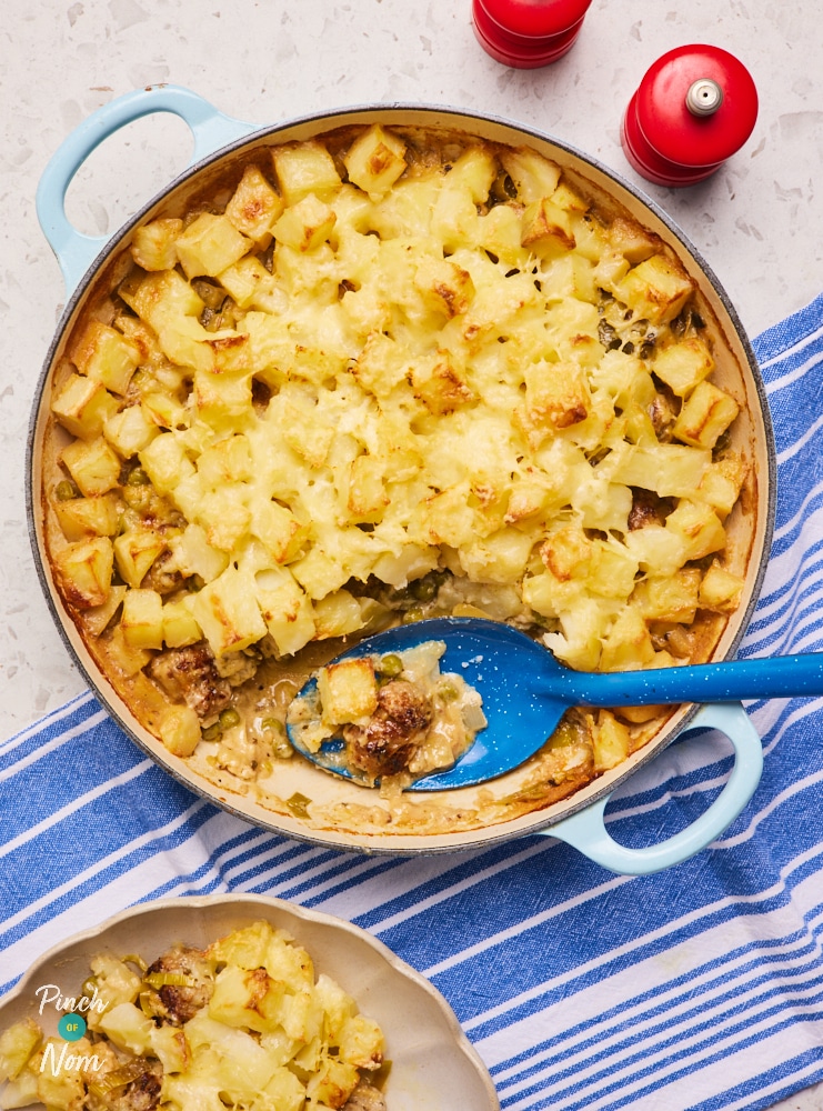 Pinch of Nom's Sausage Tartiflette is served in a pale blue, round casserole dish, placed on a blue and white striped tea towel. A deep blue serving spoon rests in the dish, where a scoop of the tartiflette has been removed and served in a small bowl nearby. The tartiflette is golden and covered in melted cheese.