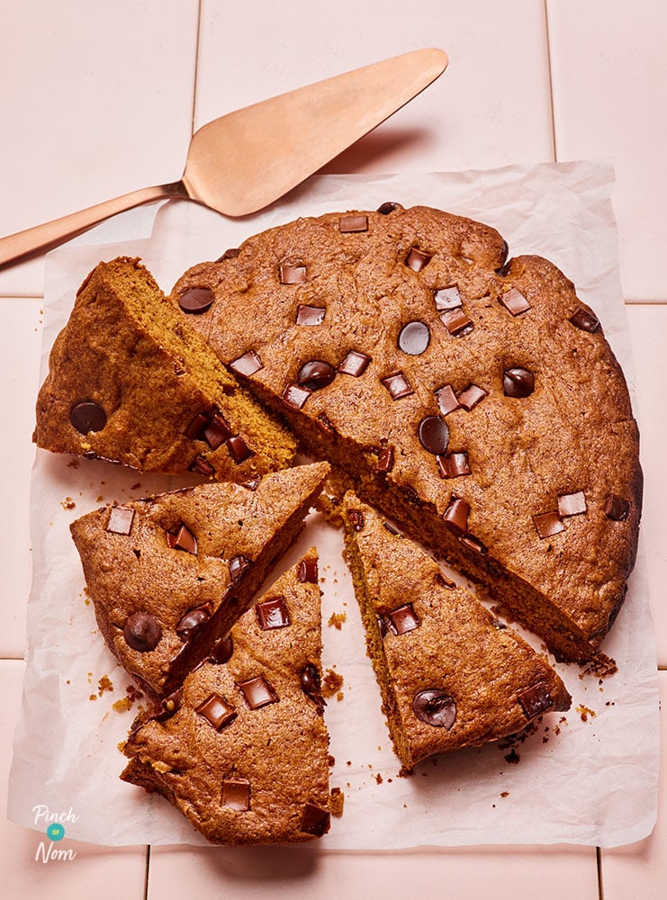 Pinch of Nom's Slow Cooker Giant Cookie is served freshly-baked on a tiled countertop; half of it has already been sliced into four portions. The gooey milk chocolate pieces are showing throughout the middle of the sponge and on top of the bake.