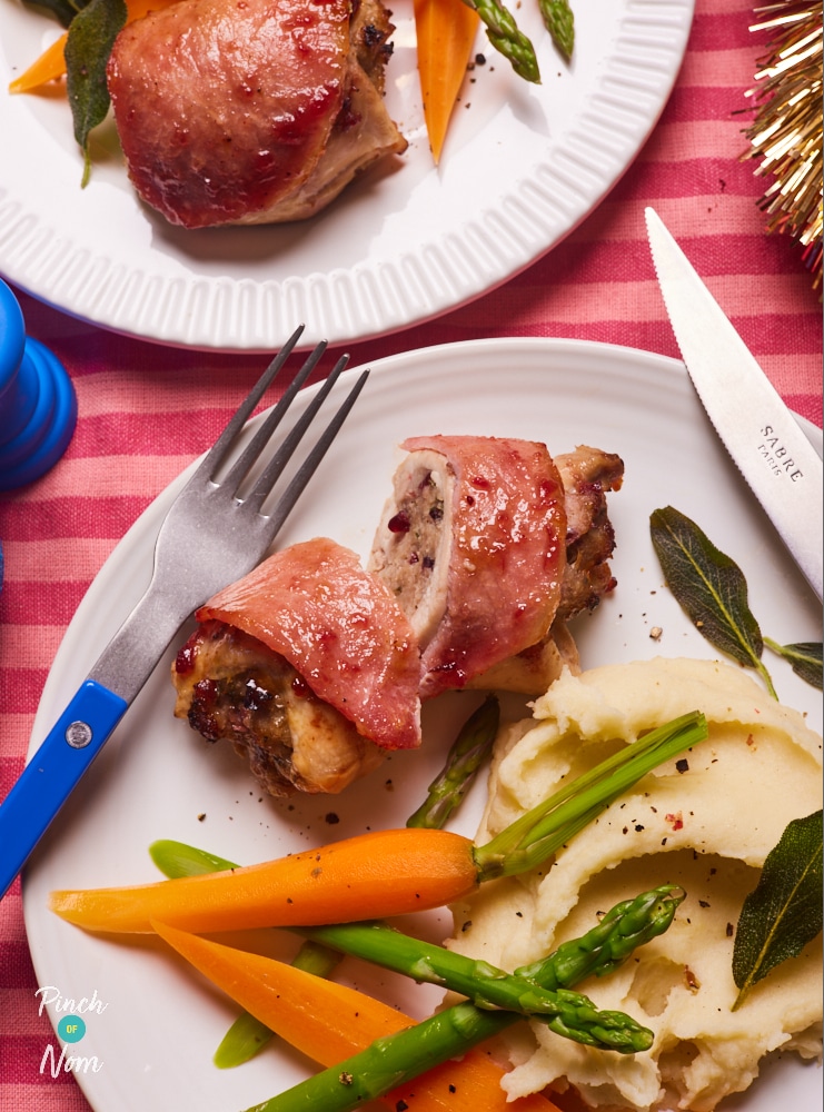 A colourful, festive table is set with two portions of Pinch of Nom's slimming-friendly Christmas Chicken Thighs recipe; the chicken thigh on the closest plate is cut open, to reveal the sausage stuffing inside. It's served with mashed potatoes and steamed veg.