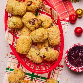 A red basket is filled with a golden, crunchy-looking batch of Pinch of Nom's slimming-friendly Christmas Dinner Croquettes, on a festive tabletop decorated with red and white chequered napkins and baubles. A pot of cranberry sauce waits to the side to be dipped.