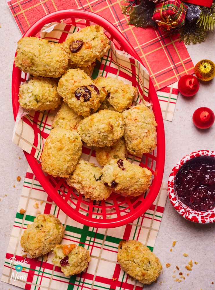 A red basket is filled with a golden, crunchy-looking batch of Pinch of Nom's slimming-friendly Christmas Dinner Croquettes, on a festive tabletop decorated with red and white chequered napkins and baubles. A pot of cranberry sauce waits to the side to be dipped.