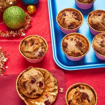 A batch of Pinch of Nom's slimming-friendly Cranberry and Orange Muffins are served on a festive bright red table, decorated with colourful baubles. One of the muffins has already been bitten into, revealing the fruity sponge centre.