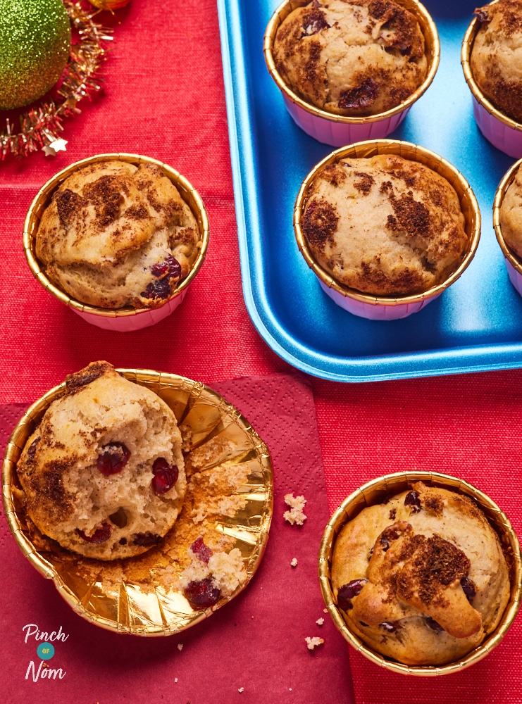 A batch of Pinch of Nom's slimming-friendly Cranberry and Orange Muffins are served on a festive bright red table, decorated with colourful baubles. One of the muffins has already been bitten into, revealing the fruity sponge centre.