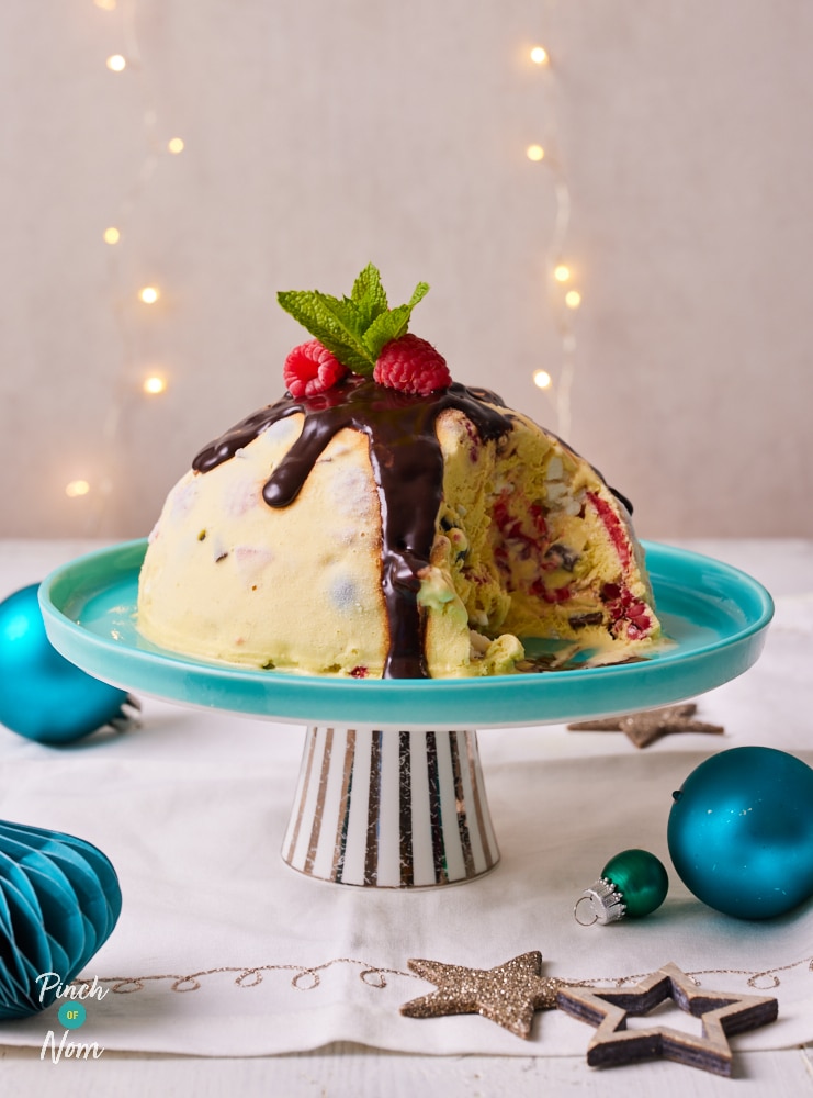 Pinch of Nom's slimming-friendly Frozen Christmas Pudding is served as the centrepiece on a festive tabletop, against a background of twinkling fairy lights. The pudding is topped with berries and a drizzle of chocolate sauce.