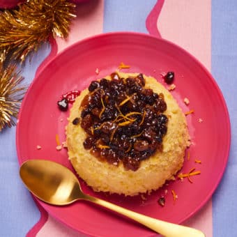 Pinch of Nom's Mince Pie Pudding is served on a red plate. The moist orange sponge is topped with a juicy layer of mincemeat. A gold spoon rests on the plate, ready to dig in.