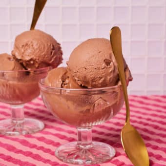 A table is placed against a light pink wall, topped with a funky-striped pink and red tablecloth. On the table, 2 portions of Pinch of Nom's low-calorie Ninja Creami Peanut Butter and Chocolate Ice Cream have been served in glass ice cream bowls. Gold-plated spoons wait to tuck in.
