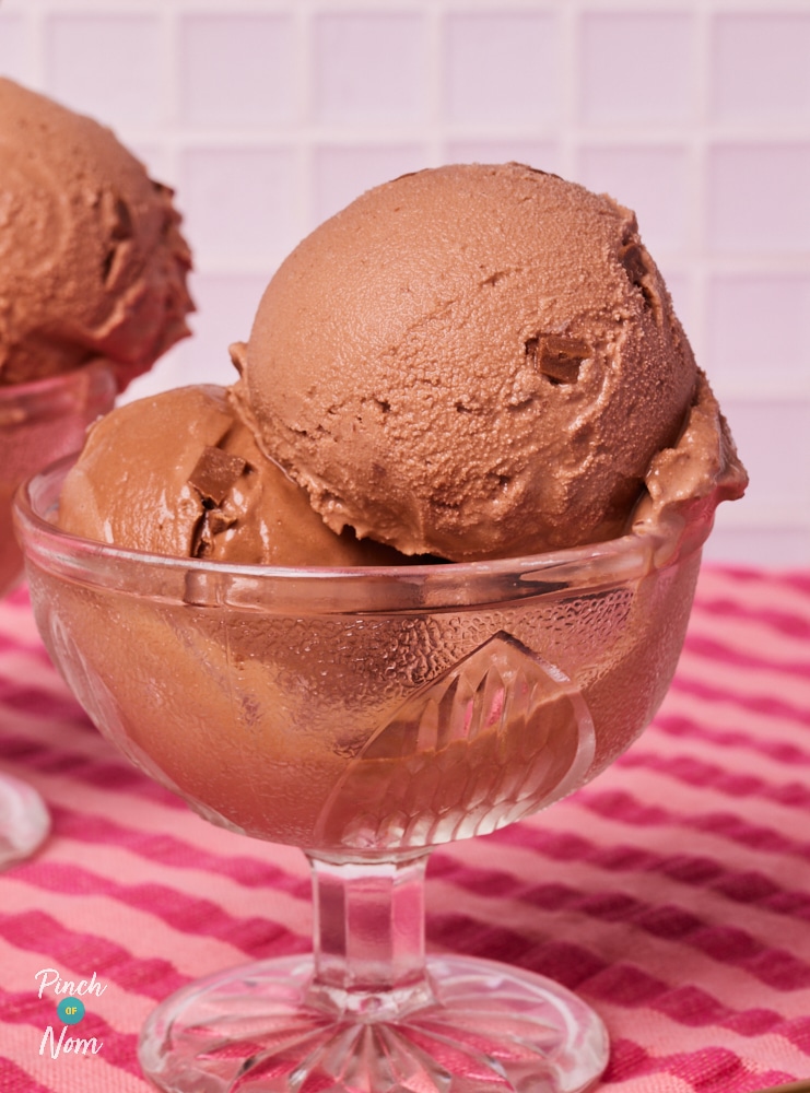 A table is placed against a light pink wall, topped with a funky-striped pink and red tablecloth. On the table, 2 portions of Pinch of Nom's low-calorie Ninja Creami Peanut Butter and Chocolate Ice Cream have been served in glass ice cream bowls. Gold-plated spoons wait to tuck in.