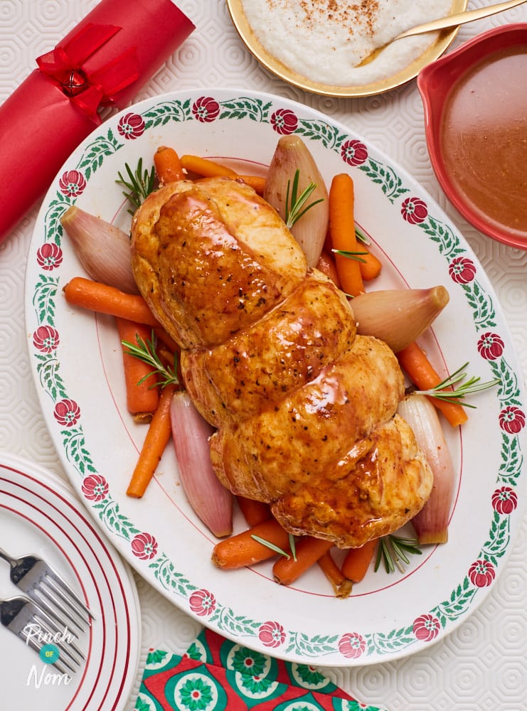 Pinch of Nom’s slimming-friendly Slow Cooker Christmas Turkey Joint is served over a bed of roast veg, on a large serving plate at the centre of a festive table.