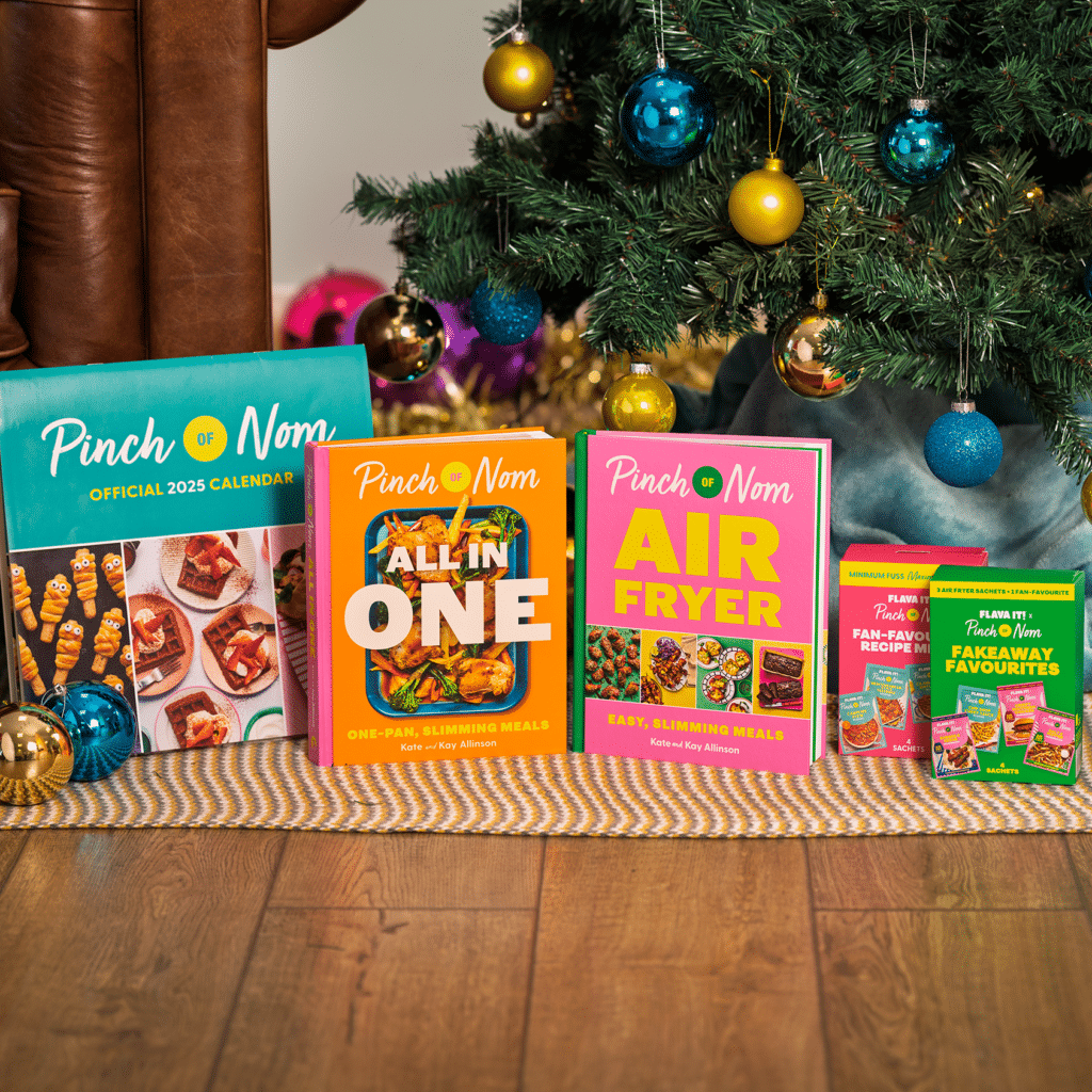 Pinch of Nom's official 2025 calendar, All in One cookbook, Air Fryer cookbook and two boxes of PON x Flava It! seasoning sachets sit below a Christmas tree.