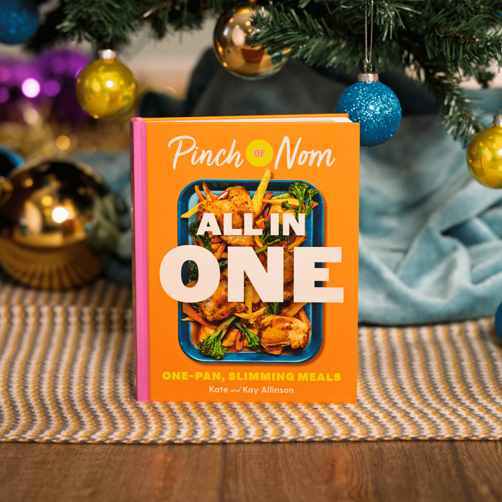 A copy of Pinch of Nom: All in One stands under a colourfully decorated Christmas tree, as though it has just been unwrapped on Christmas morning.